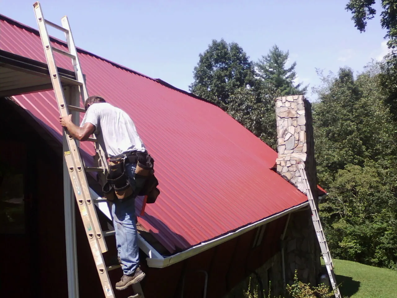 Roofer on a ladder beside a red metal roof and stone chimney — Soffit & Fascia Repair by Roofing Brothers in Harwood